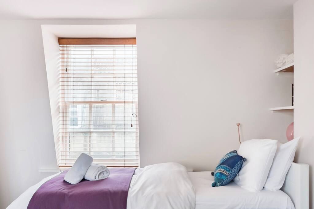 Ein Schlafzimmer mit einem Bett, einem Fenster mit Jalousien, einer violetten Decke und einem weißen Kissen.