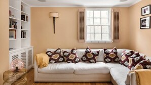 A living room with a beige sofa, patterned cushions, a white bookshelf, and a window with shutters. - Murphy's House Central Townhouse W/courtyard (Bath)