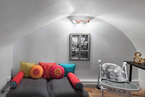 Een compacte kamer met een schuine kap, een bed met beddengoed met een patroon, kleurrijke kussens, een klein raam met uitzicht en een baby schommel.