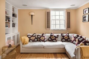 A living room with a beige sofa, patterned cushions, a white bookshelf, and a window with shutters. - Murphy's House Central Townhouse W/courtyard (Bath)