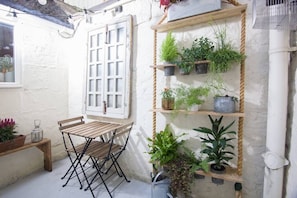 A small outdoor seating area with a wooden table and chairs, surrounded by potted plants on shelves and hanging from the wall.