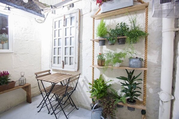 A small outdoor seating area with a wooden table and chairs, surrounded by potted plants on shelves and hanging from the wall.
