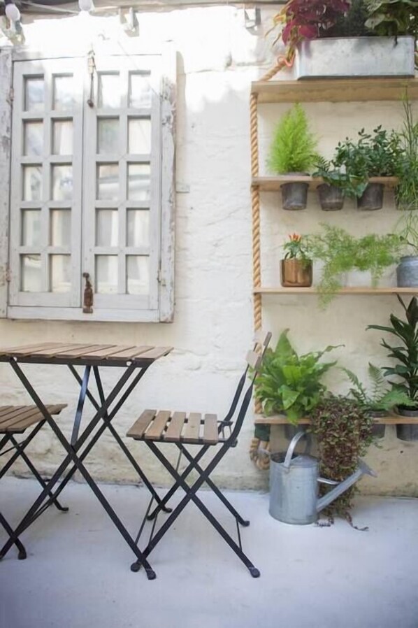 A rustic outdoor seating area with a wooden table and metal chairs, a white wall with a window, and shelves with potted plants.