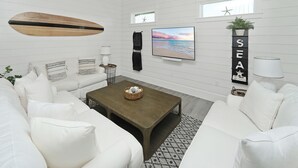 Un salon moderne avec un ensemble de canapés blancs, une table basse en bois, un téléviseur à écran plat et une étagère murale agrémentée d’objets décoratifs.