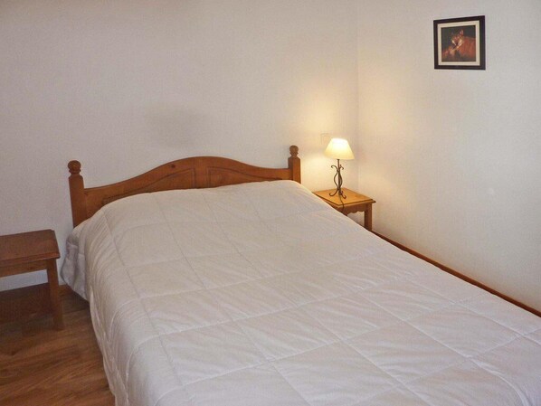 A single bed with a wooden headboard and a white quilted bedspread in a room with a wooden floor. There is a bedside table with a lamp and a framed picture on the wall.