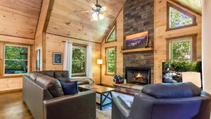 A log cabin interior with a fireplace, leather sofas, wooden walls, and large windows. - Spacious, New Cabin! New Hot Tub, Fire Pit, and Game Room! Dog-Friendly! (Blue Ridge)