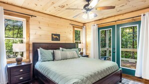 A wooden cabin bedroom with a bed, nightstands, a ceiling fan, and a view of the outdoors. - Spacious, New Cabin! New Hot Tub, Fire Pit, and Game Room! Dog-Friendly! (Blue Ridge)