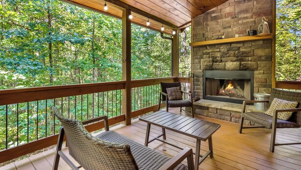 A covered outdoor deck with a stone fireplace, wicker furniture, and a view of the forest. - Spacious, New Cabin! New Hot Tub, Fire Pit, and Game Room! Dog-Friendly! (Blue Ridge)
