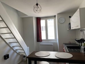 Une cuisine avec une table à manger, une fenêtre avec un rideau, une horloge et un escalier.