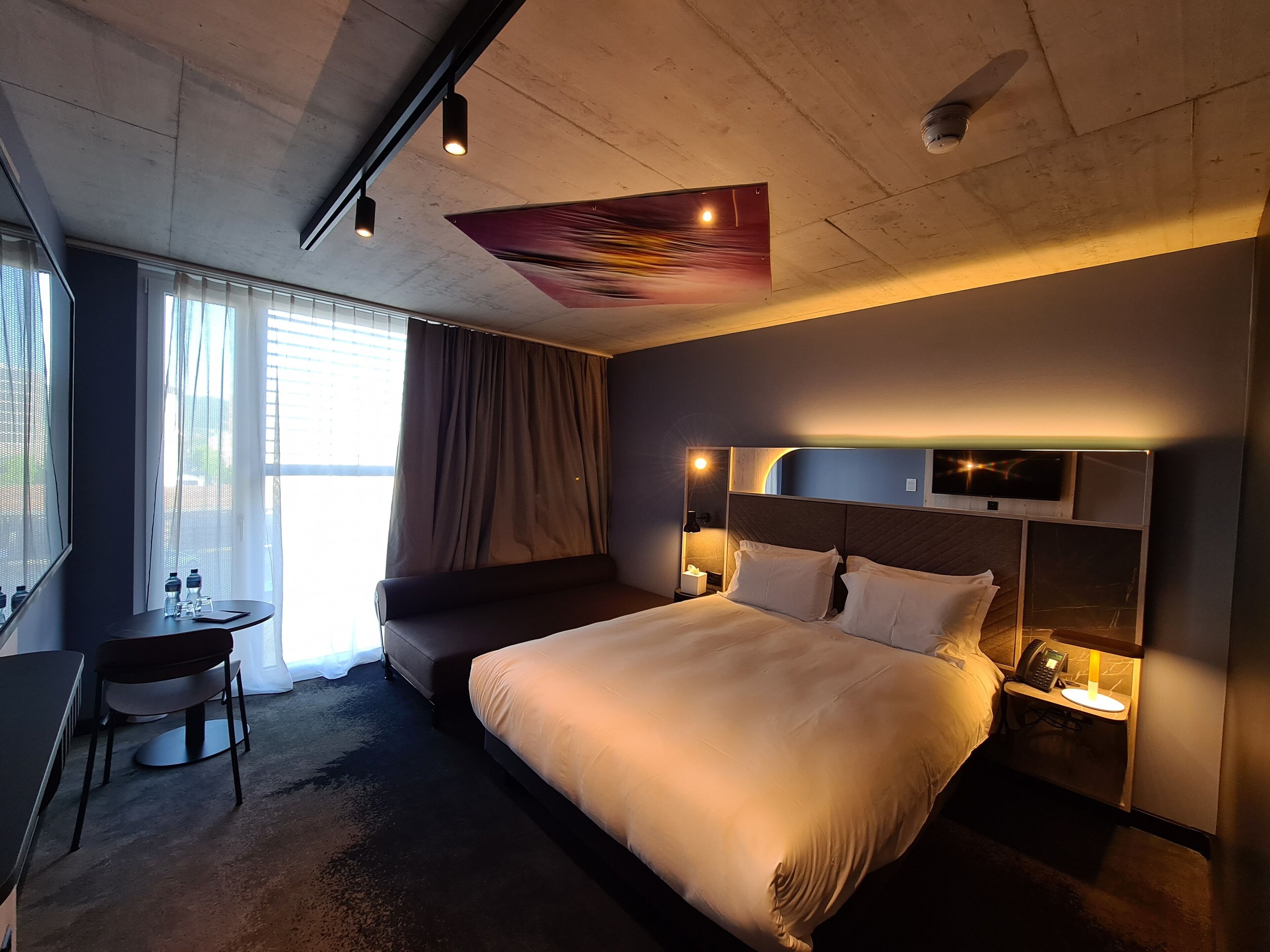 Habitación de hotel moderna con una cama grande, mesita de noche con lámpara y un escritorio con silla.