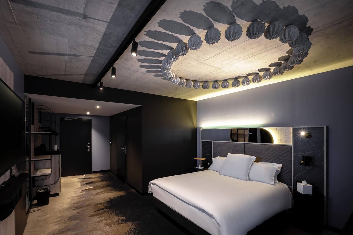 A modern hotel room with a large bed, a ceiling with a unique light fixture, and a wall-mounted TV.