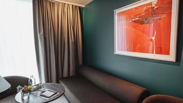 Une chambre d’hôtel moderne avec un canapé, une petite table ronde ornée d’un vase de fleurs, un grand tableau abstrait accroché au mur et une fenêtre avec des rideaux.