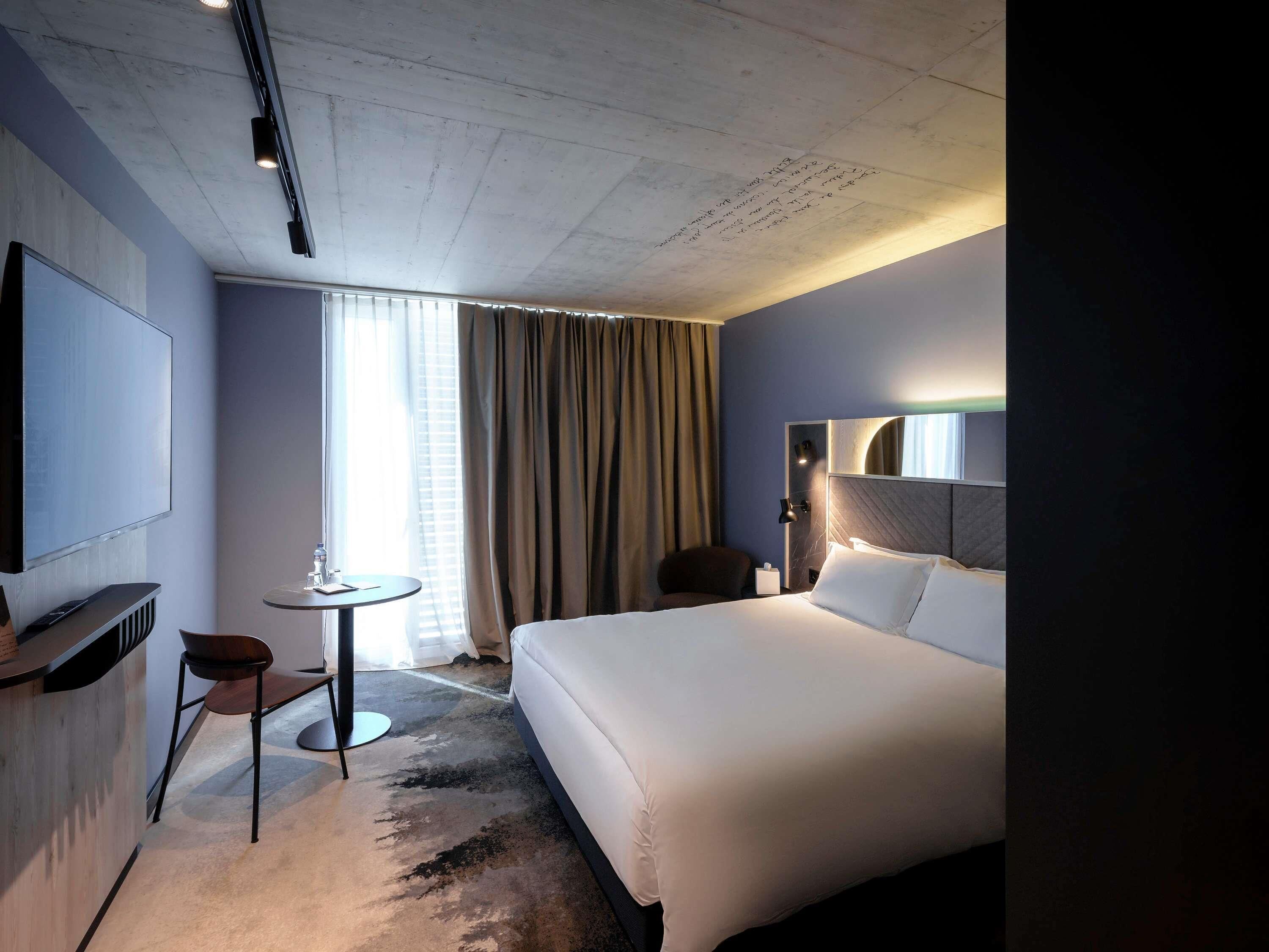 A hotel room with a bed, a desk with a chair, a TV, and a window with curtains.