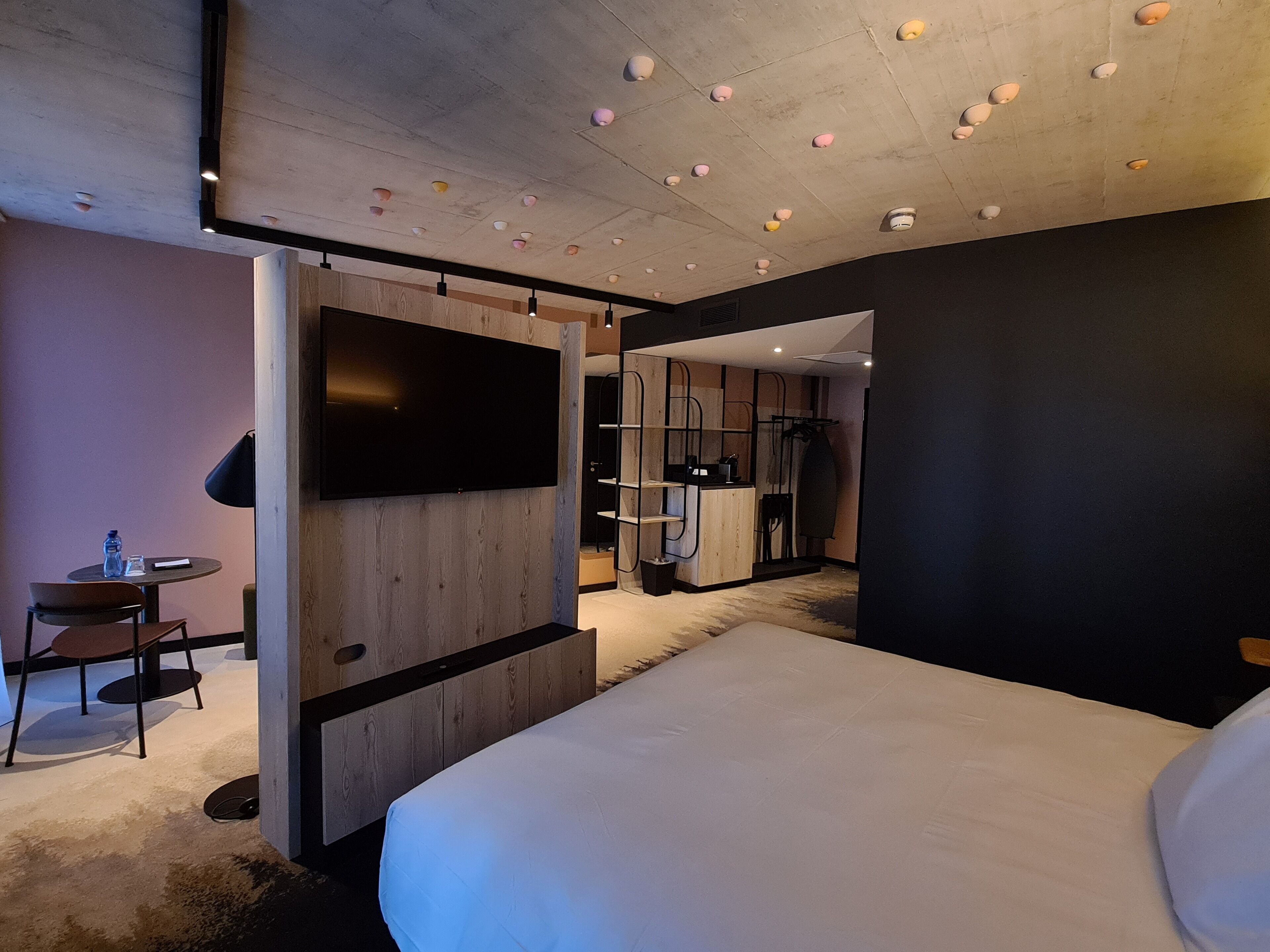 Ein modernes Hotelzimmer mit einem großen Bett, einem flachbildfernseher an der Wand, einem kleinen Tisch mit Stuhl und einer Küchenzeile.