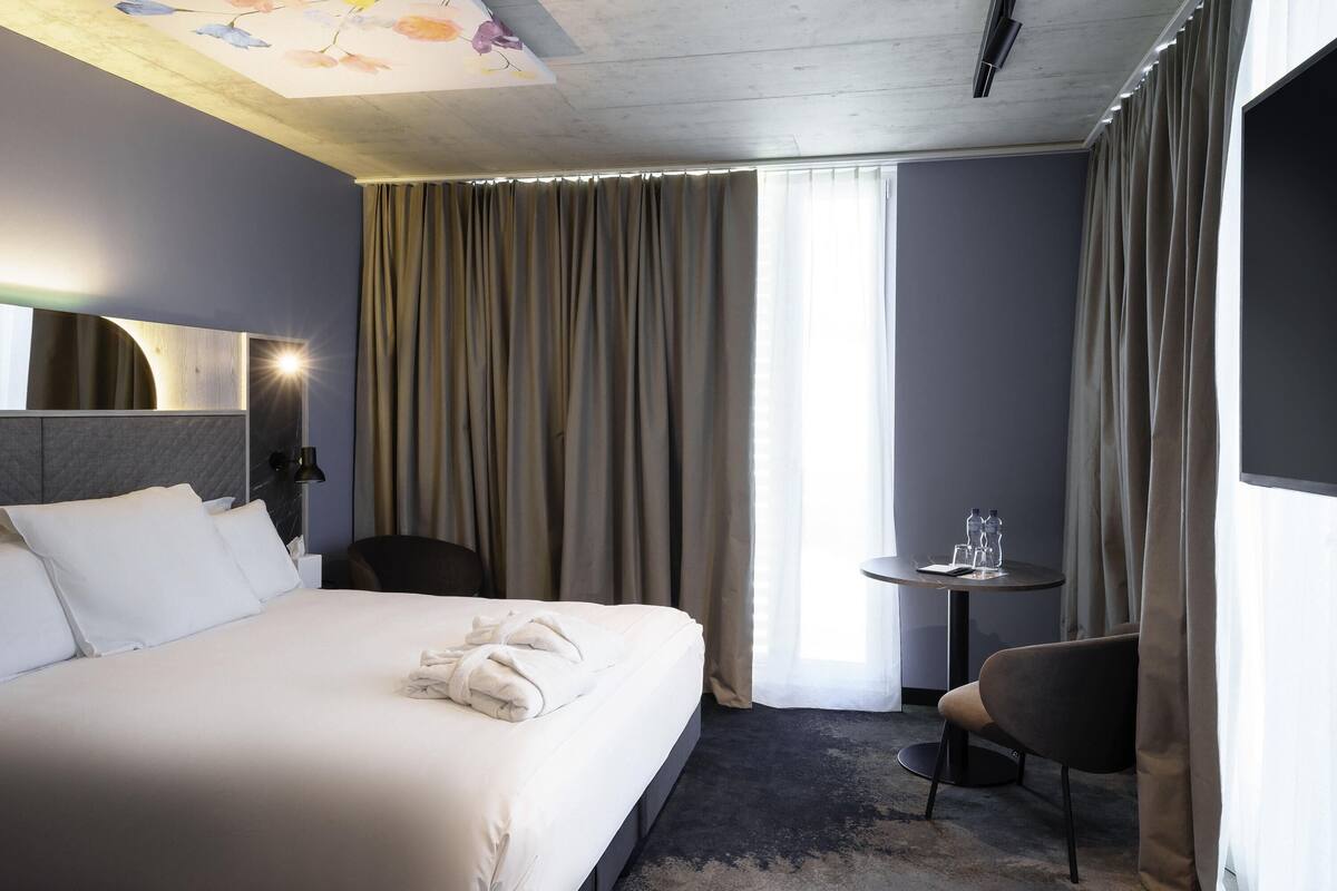 A hotel room with a large bed, a small table with water bottles, a chair, and a TV on the wall.