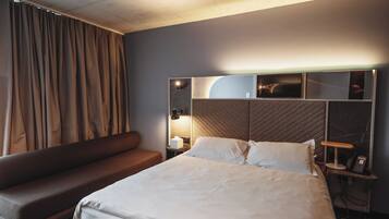 Habitación de hotel moderna con una cama grande, un sofá, una mesita de noche con lámpara y un televisor montado en la pared.