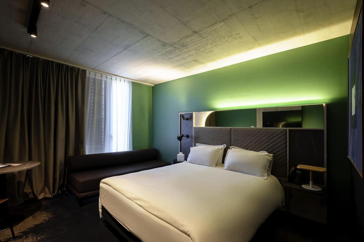 A hotel room with a large bed, a bedside table, a lamp, a window with curtains, and a green accent wall.