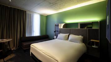 A hotel room with a large bed, a bedside table, a lamp, a window with curtains, and a green accent wall.