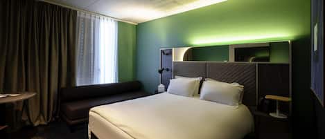 A hotel room with a large bed, a bedside table, a lamp, a window with curtains, and a green accent wall.
