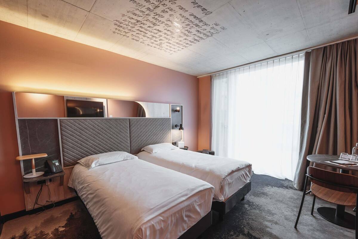 A hotel room with two beds, a desk, a chair, and a large window with curtains.