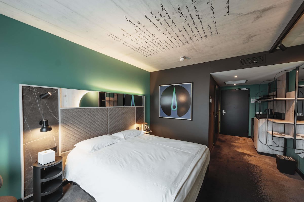 A modern hotel room with a large bed, a desk with a lamp, a wall-mounted artwork, and a staircase leading to an upper level.
