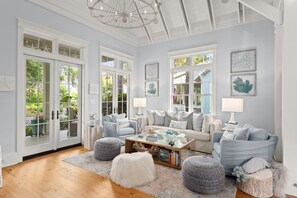 A spacious living room with a large window, a chandelier, and a cozy seating area with a coffee table and ottomans.