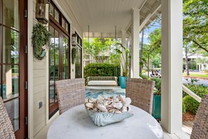 A covered porch with wicker furniture, a table with seashells, and a view of a pool and greenery.
