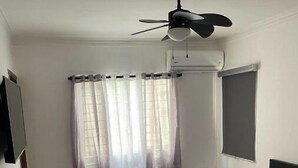 Une chambre à coucher comprenant un lit, un ventilateur de plafond, une fenêtre avec des rideaux, une table de chevet avec une lampe et un téléviseur fixé au mur.