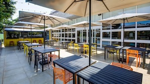 Ein Außenbereich mit Terrasse, Tischen, Stühlen und großen Sonnenschirmen, im Hintergrund ein Glasgebäude.