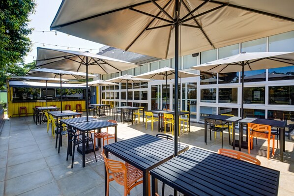 Un patio al aire libre con mesas y sillas, sombrillas grandes y un edificio de vidrio al fondo.