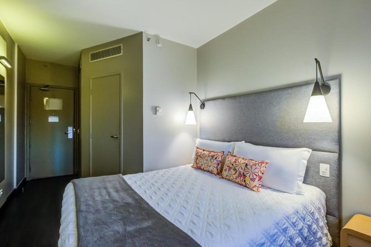 A hotel room with a bed, two wall-mounted lamps, a door, and a carpeted floor.