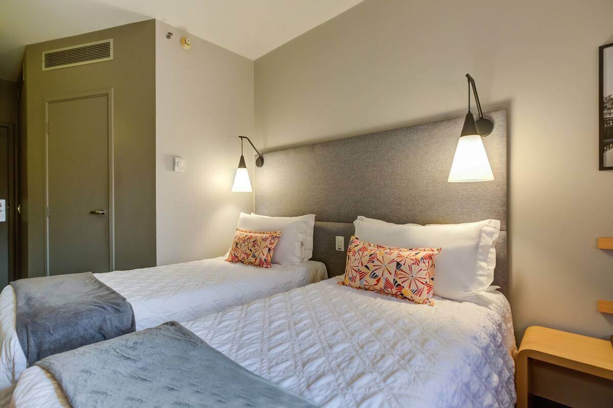 Une chambre d’hôtel avec deux lits, chacun recouvert d’une courtepointe blanche matelassée et agrémenté de coussins décoratifs. Des lampes de lecture sont fixées au mur au-dessus de chaque lit.