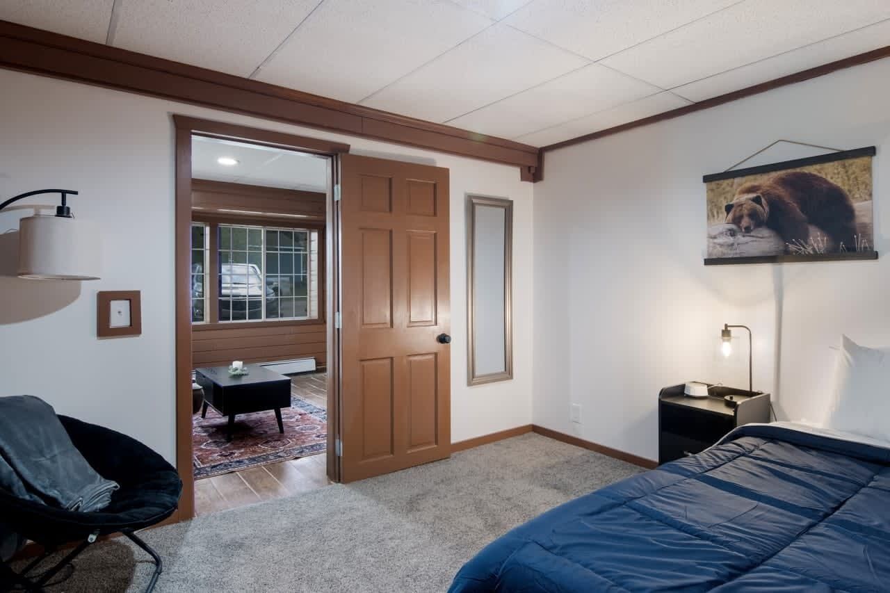 Un dormitorio con cama, una silla, una mesita de noche, una puerta y un cuadro colgado en la pared.
