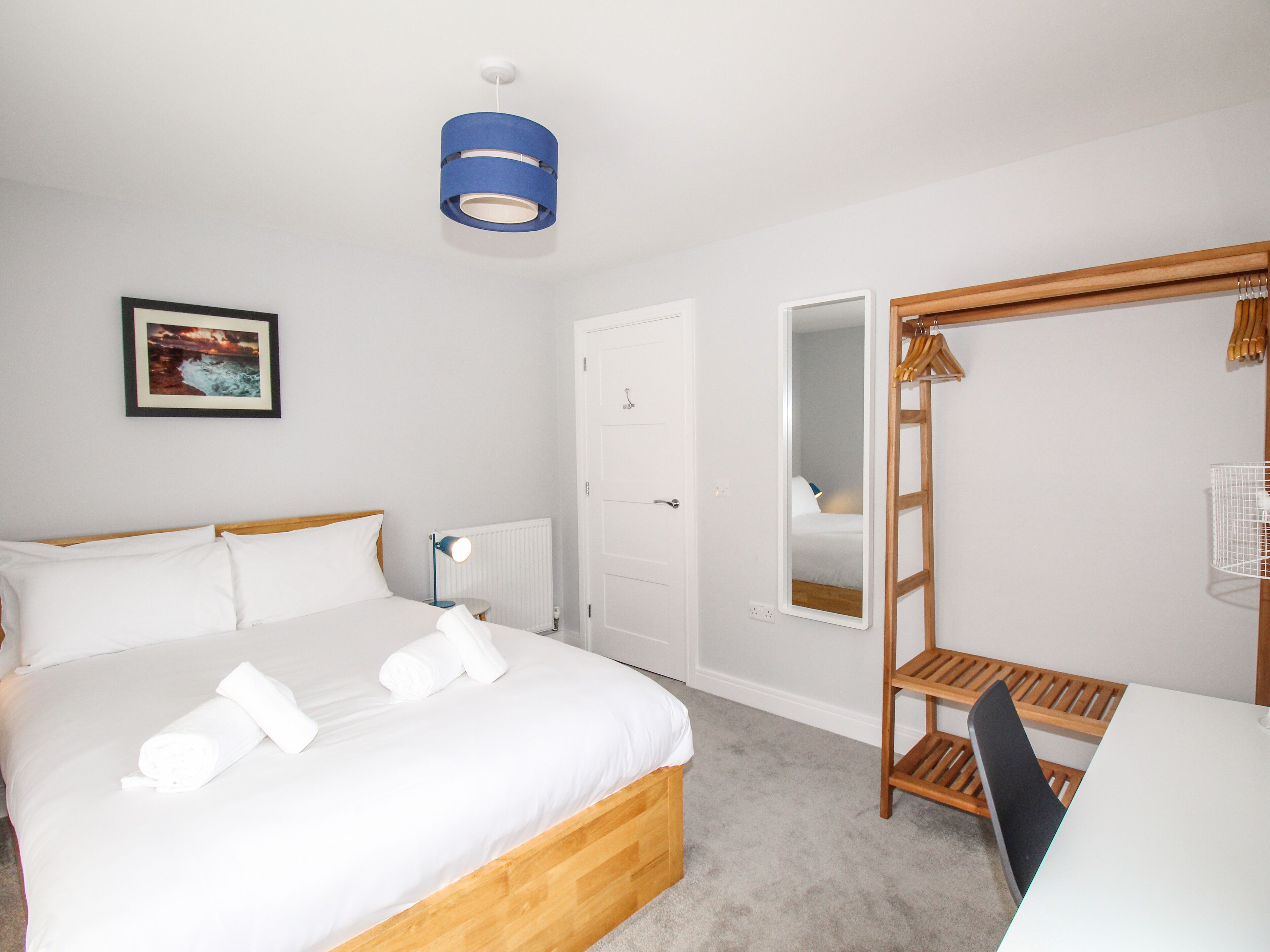 A bedroom with a bed, a bedside lamp, a mirror, a wooden chair, and a hanging coat rack.