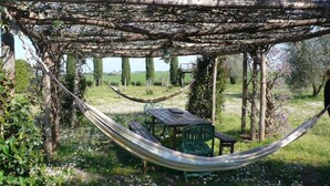 A hammock under a pergola with a table and chairs, surrounded by greenery and flowering plants.