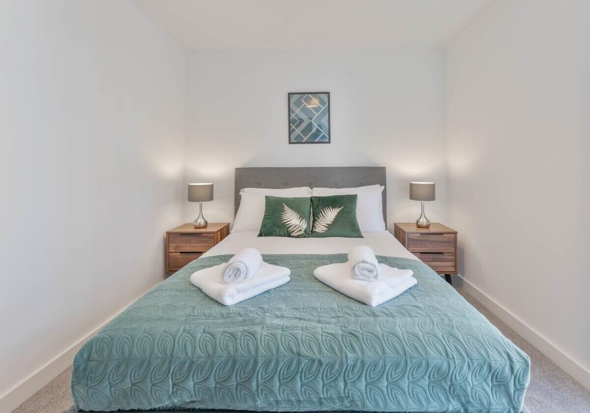 A neatly made bed with a green bedspread, two pillows, and two rolled towels. There are two bedside tables with lamps.