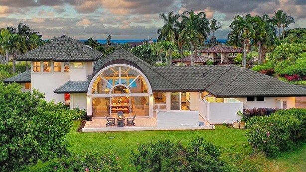 A modern house with a large glass bay window, surrounded by lush greenery and palm trees.