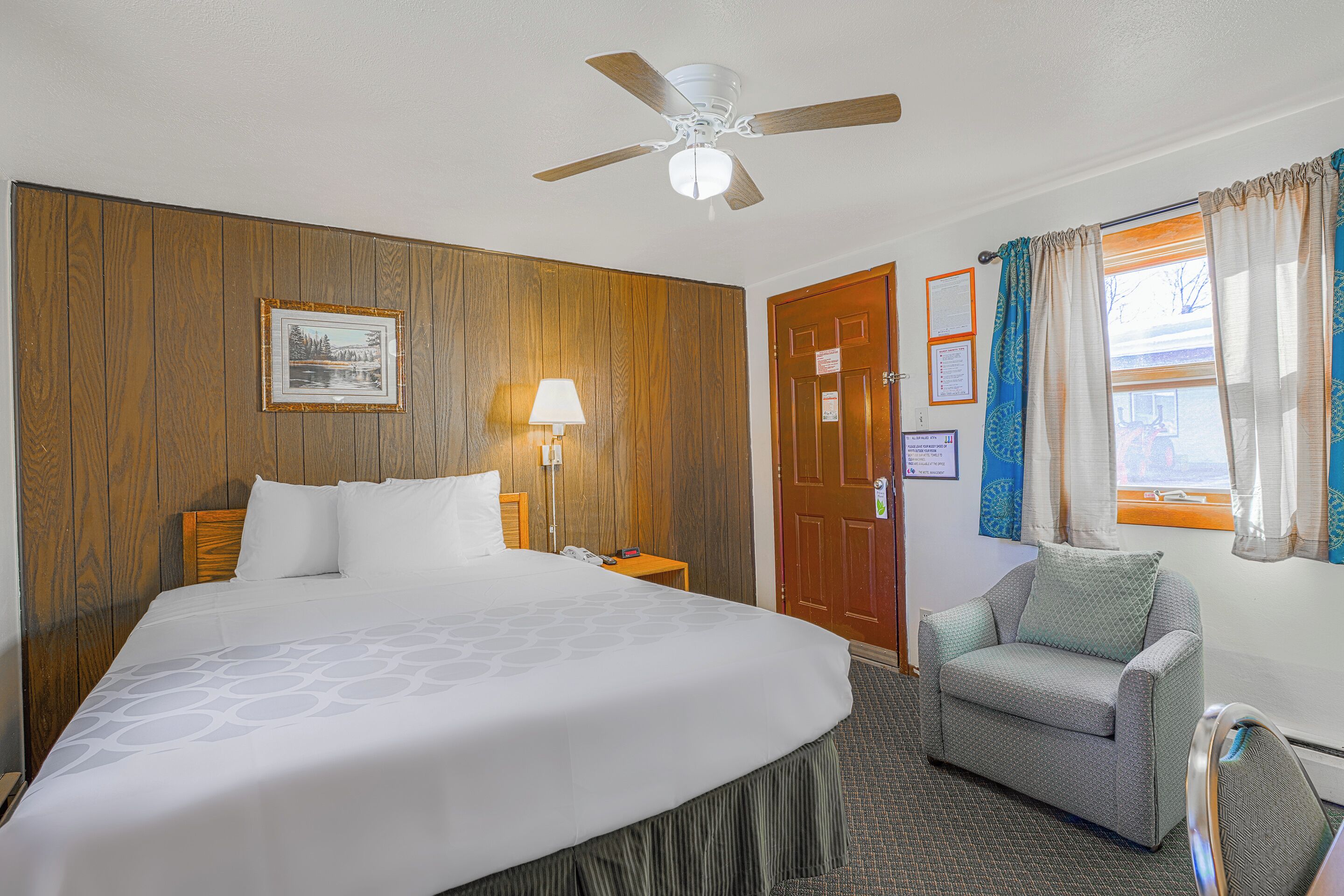 Une chambre d’hôtel avec un grand lit, une chaise, un bureau, un ventilateur de plafond, une lampe et une fenêtre avec des rideaux.