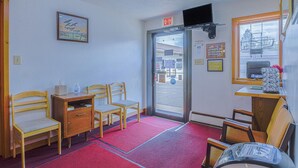 Een hotel lobby met houten stoelen, een klein houten bureau, een rode loper en een deur met een nooduitgangsbord.