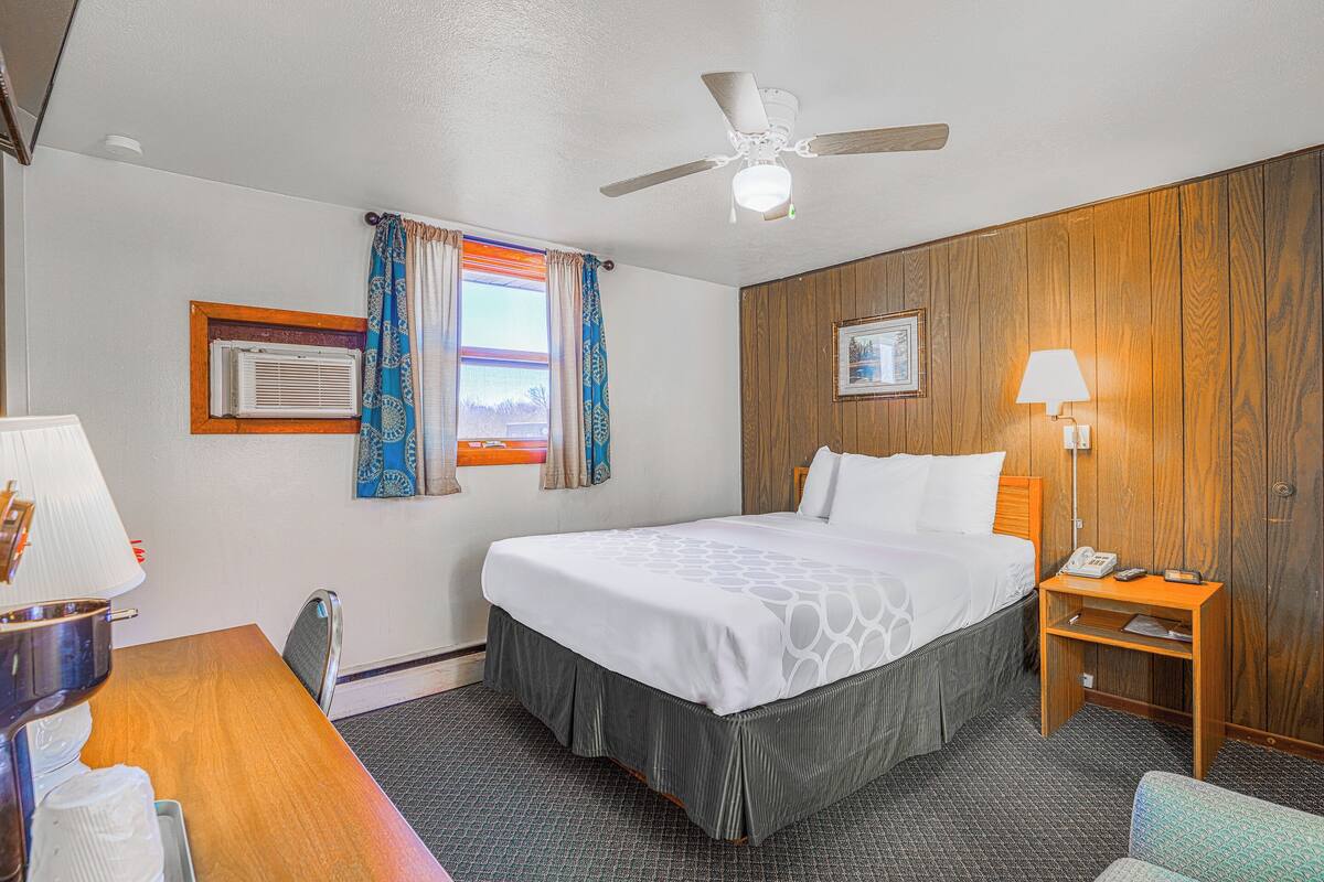 Hotelkamer met een bed, bureau, stoel, nachtkastje, lamp, plafondventilator, raam met gordijnen en een lijst met een afbeelding aan de muur.