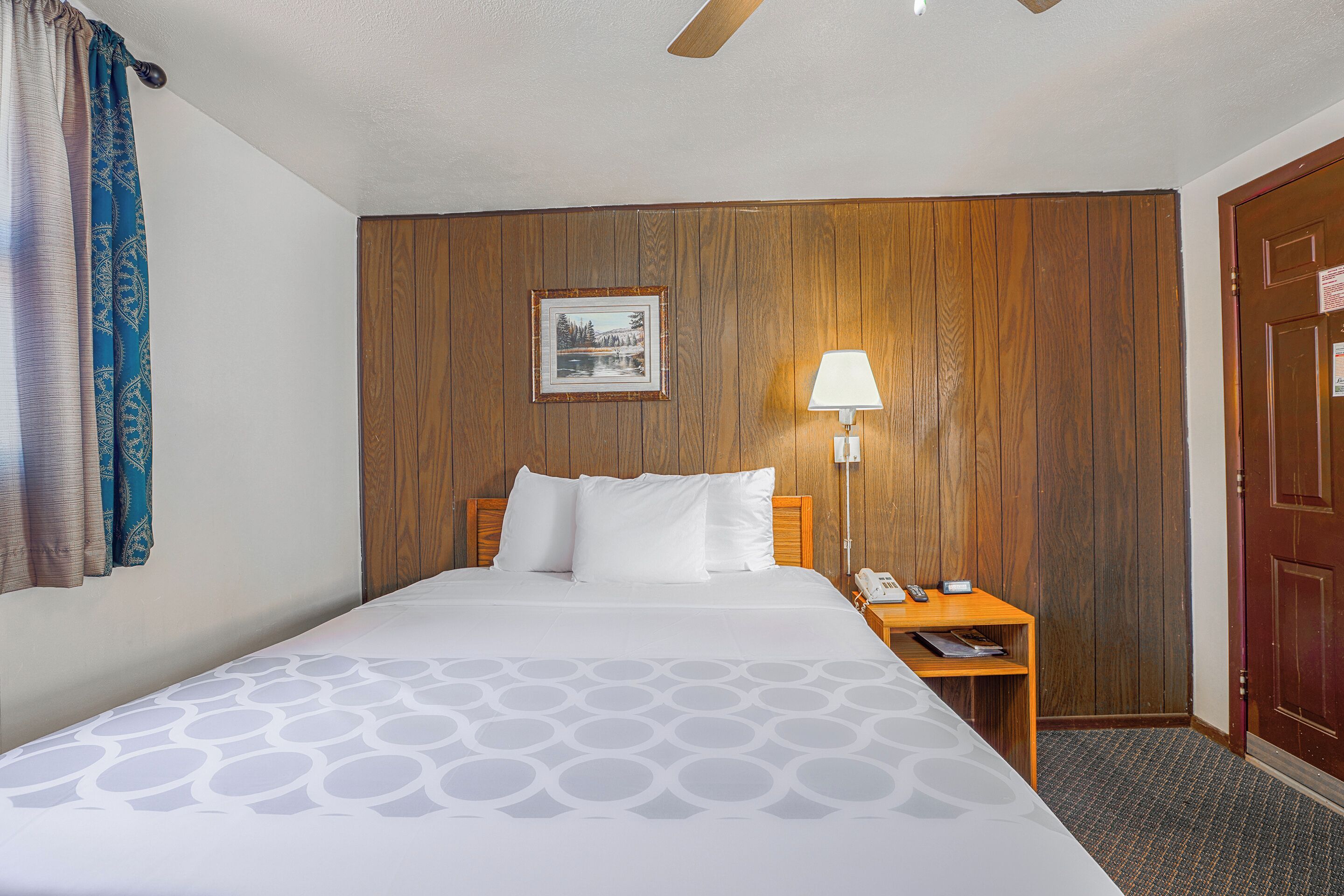 A hotel room with a bed, a nightstand with a lamp, a wooden wall panel, and a door.