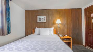 A hotel room with a bed, a nightstand with a lamp, a wooden wall panel, and a door.
