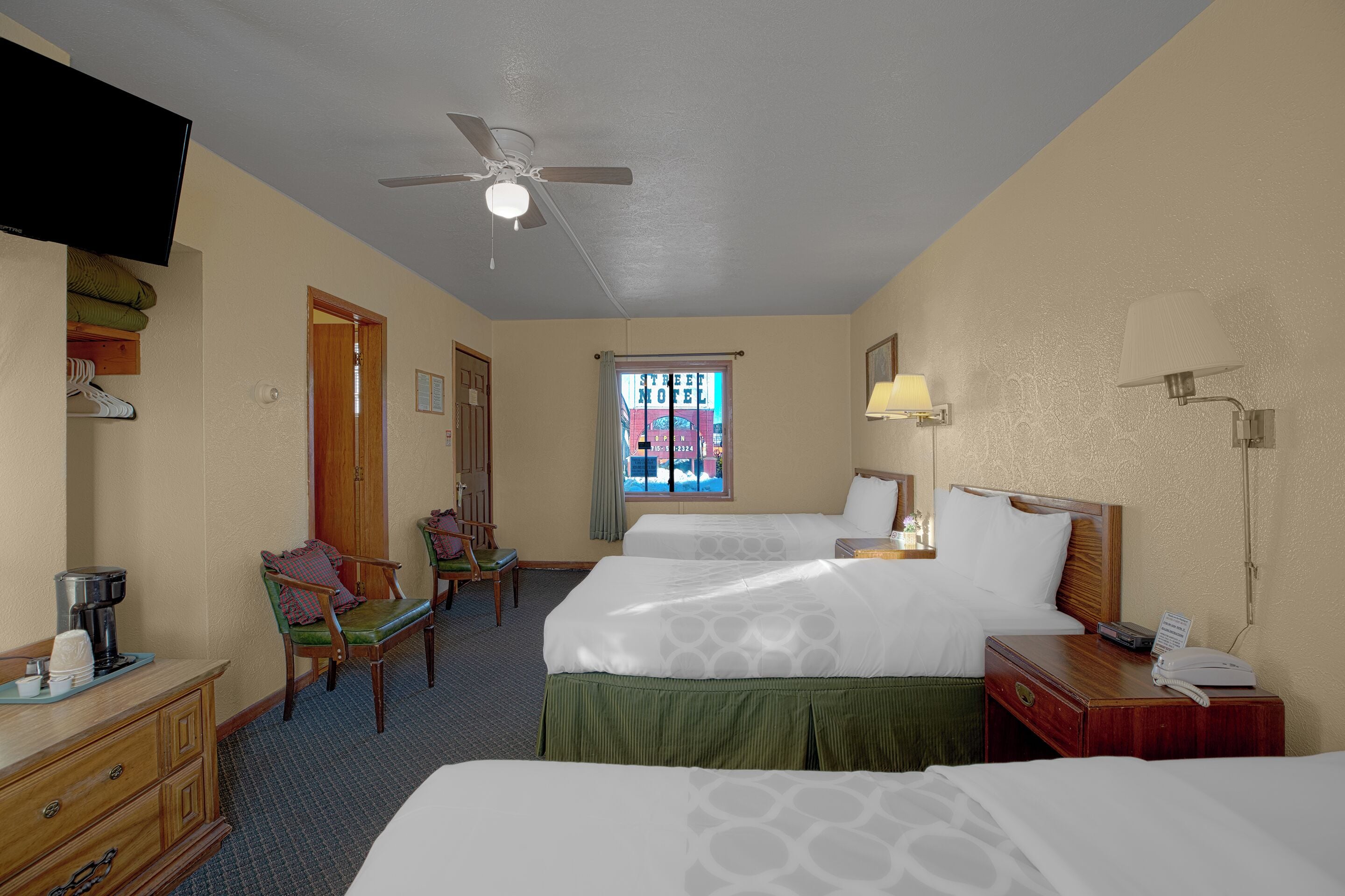 A hotel room with two beds, a desk, a chair, a television, a ceiling fan, and a window with a view of a building.