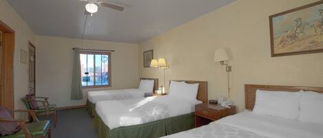 Een hotelkamer met twee bedden, een plafondventilator, een raam met gordijnen, een bureau met een lamp en een schilderij aan de muur.