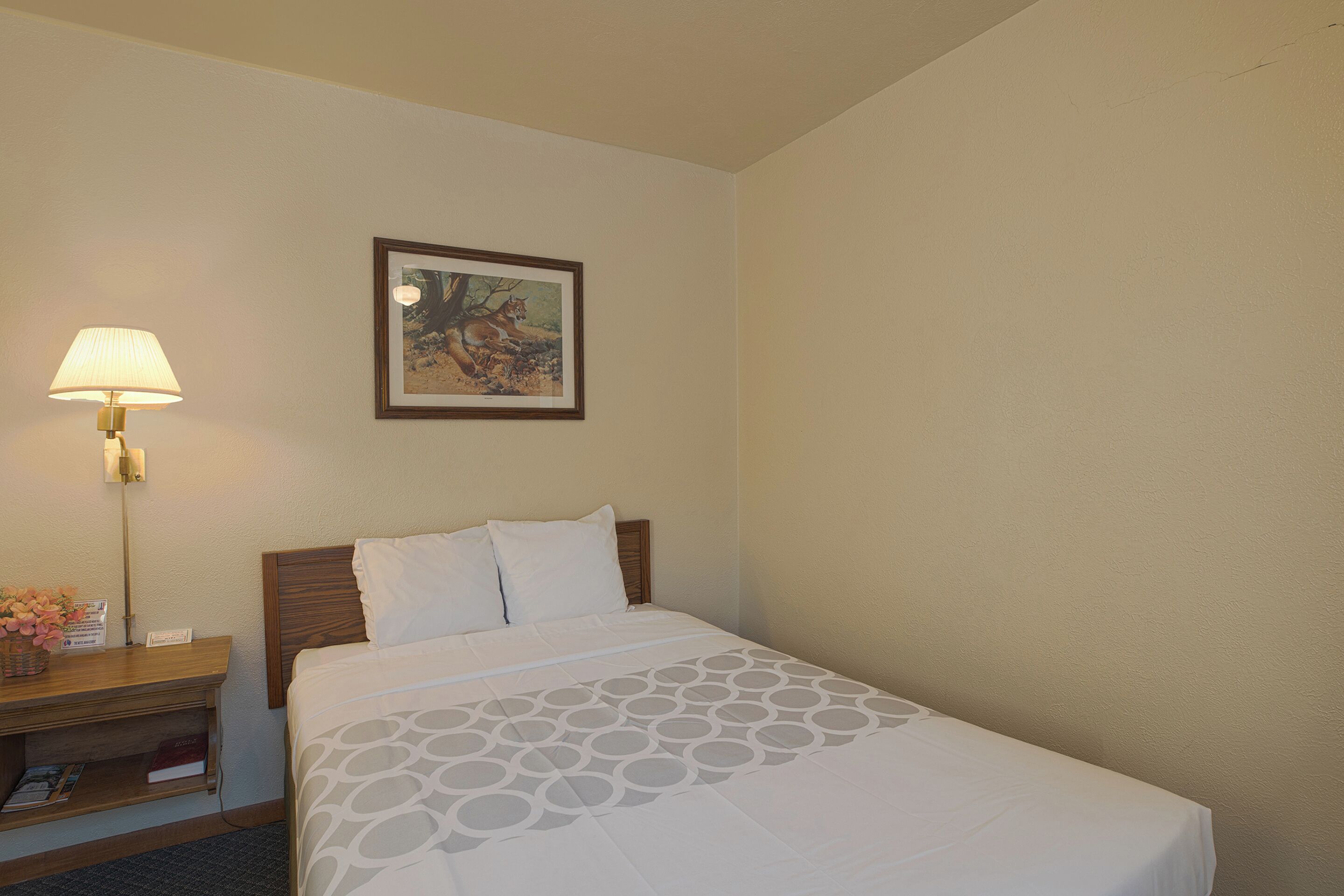 A hotel room with a neatly made bed, a bedside table with a lamp, a framed picture on the wall, and a small basket with flowers.