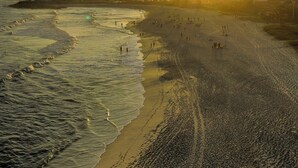 A beach with footprints leading to the water, a distant hill with buildings, and the sun setting over the horizon. -  Viver Pousada (Saquarema)