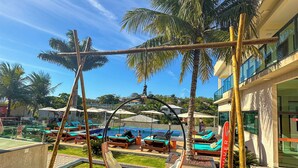 A pool area with lounge chairs and a swing chair, surrounded by palm trees and a building with a sign. -  Viver Pousada (Saquarema)