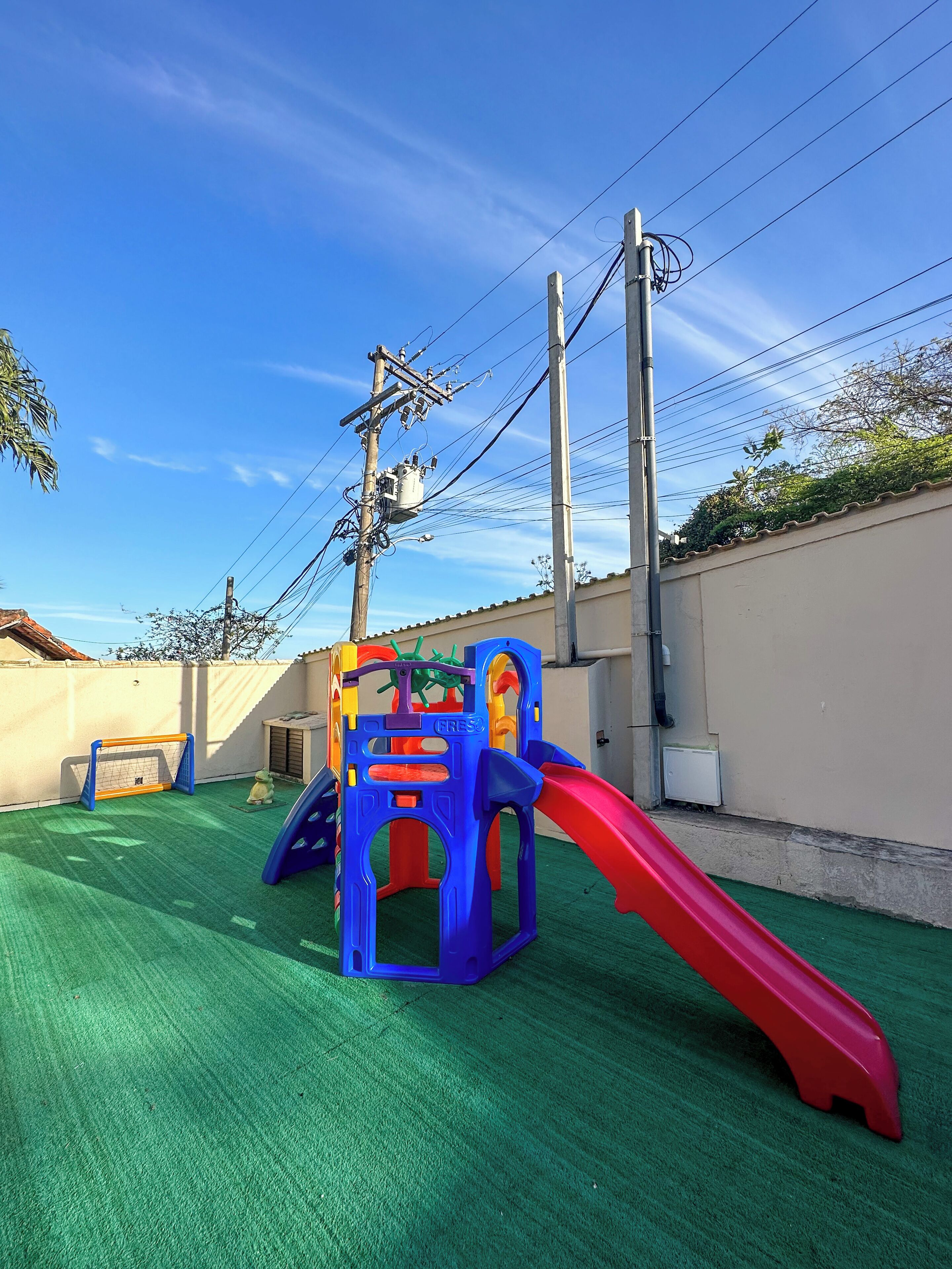 A playground with a slide and climbing structures, surrounded by a green turf area and a beige wall.