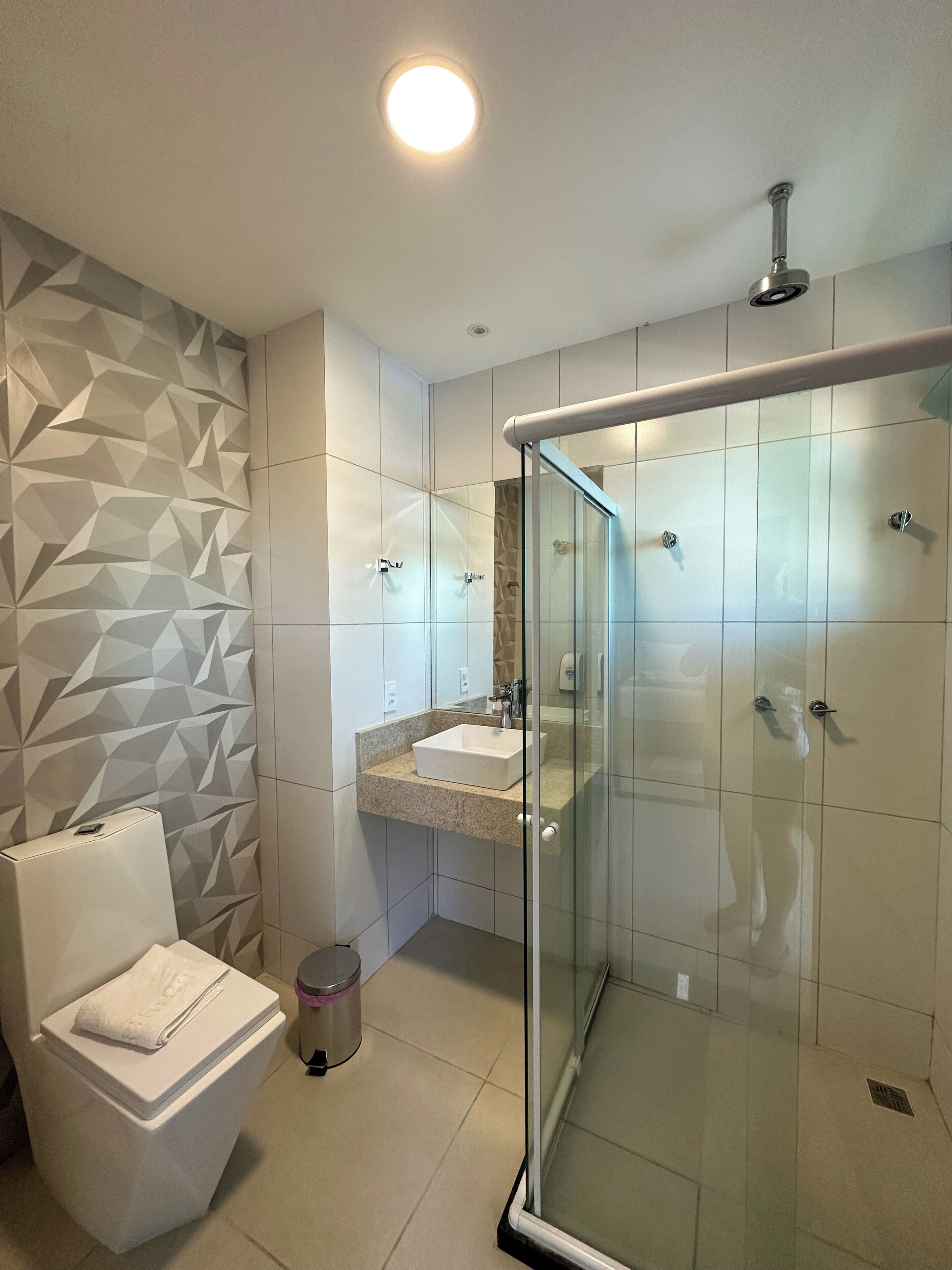 Un baño moderno con pared de estampado geométrico, ducha con cerramiento de vidrio, lavabo blanco y un inodoro con una toalla encima.