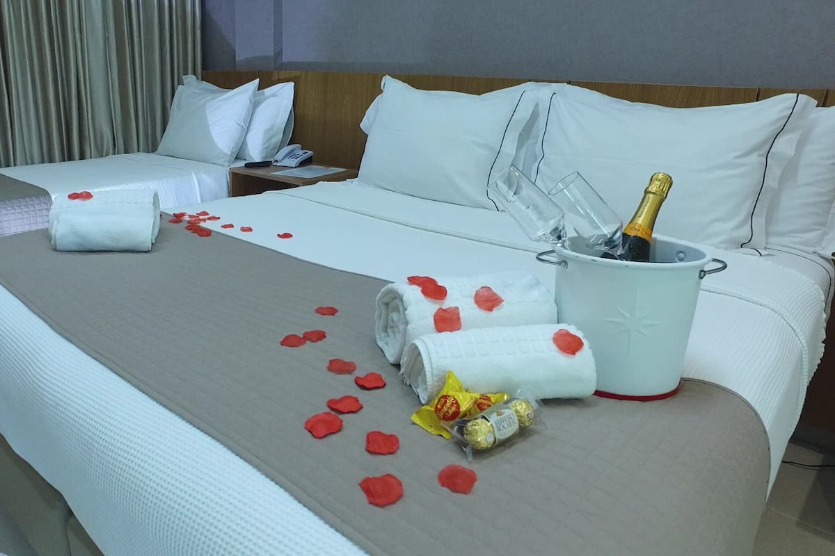 A hotel room with a bed, a nightstand, a bedside table, a bottle of champagne, and rose petals on the bed.